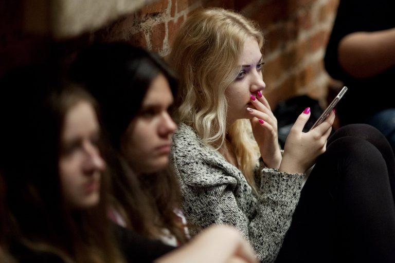 ALT - Twarze trzech nastolatek. Jedna z nich trzyma przed twarzą smartfon, drugą dłonią zasłania usta.