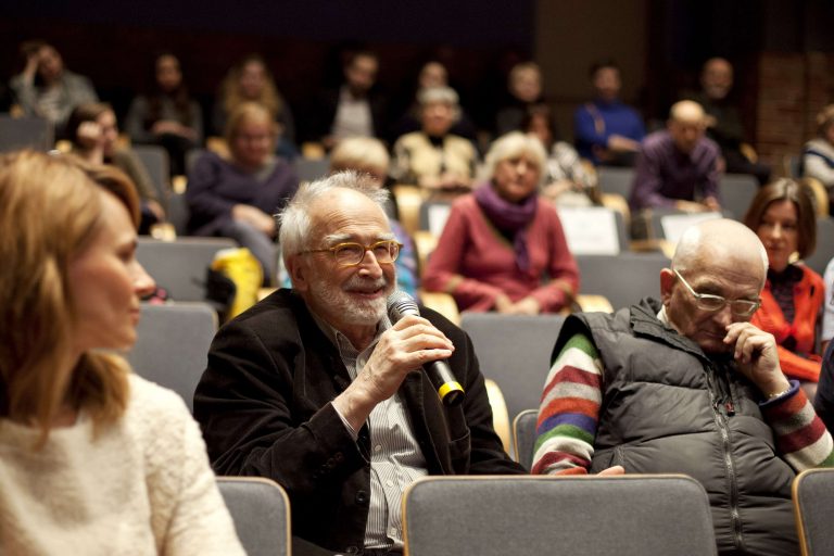 ALT - Siwy, uśmiechnięty mężczyzna w okularach siedzi wśród publiczności. W prawej dłoni trzyma mikrofon.