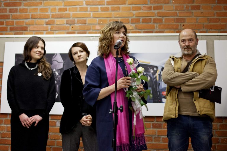 ALT - Kobieta stoi przed mikrofonem. W dłoniach kwiaty. Z tyłu dwie kobiety i mężczyzna. Spoglądają na mówiącą.