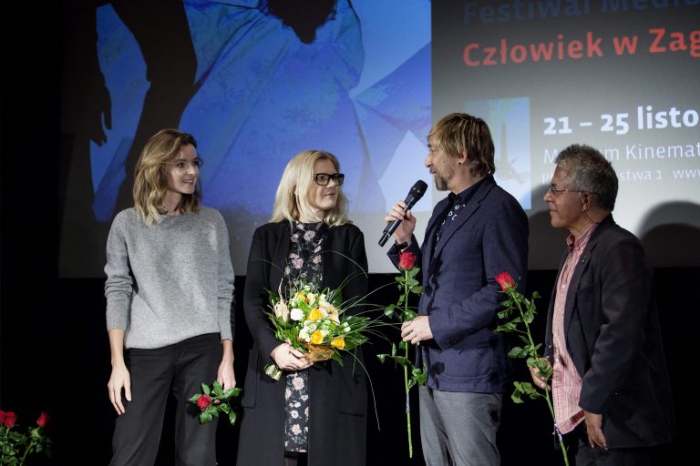 ALT - Dwie kobiety i dwóch mężczyzn stoją w rzędzie. Mężczyzna trzyma mikrofon, kobieta kwiaty.