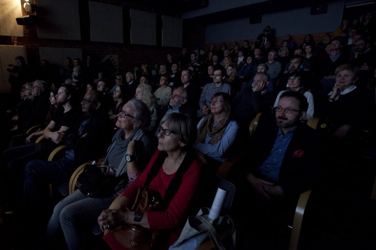 ALT - Sala kinowa z wyłączonym oświetleniem. Słabo widoczna grupa kilkudziesięciu osób. Patrzą na wprost.