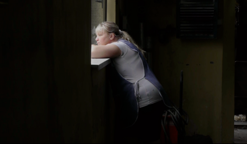 ALT - Młoda kobieta opiera się łokciami o parapet okna. Wygląda na zewnątrz. Widok z boku.