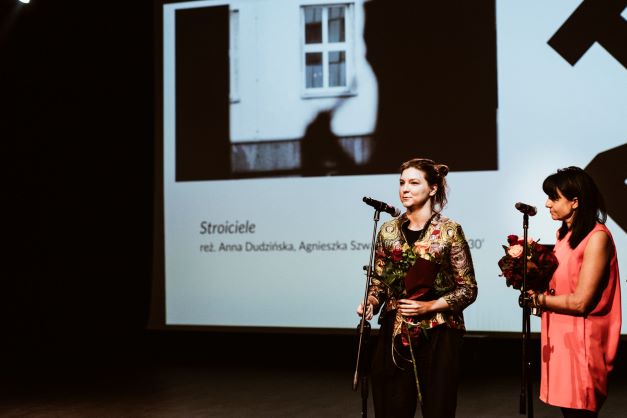 ALT - Dwie młode kobiety stoją przy mikrofonach. W tle ekran ze zdjęciem fasady budynku i napisem stroiciele.