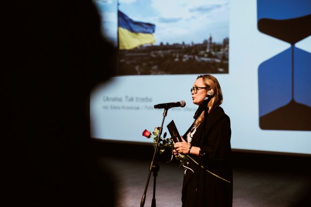 ALT - Kobieta w średnim wieku stoi przed mikrofonem. W tle ekran ze zdjęciem panoramy miasta i flagą Ukrainy.