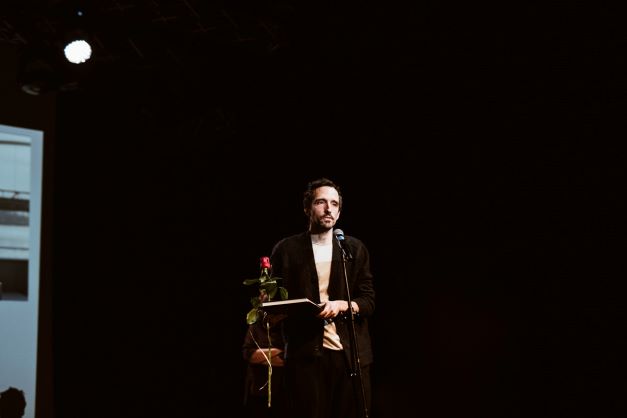 ALT - Młody mężczyzna w marynarce stoi przed mikrofonem, trzyma różę i dyplom.