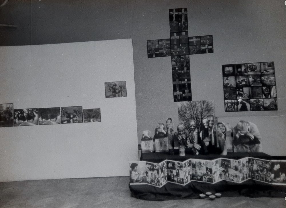 ALT - Ściana w pokoju. Główny element wiszący na ścianie to krzyż. Obok fotografie. Pod krzyżem stoją na stoliku figurki.