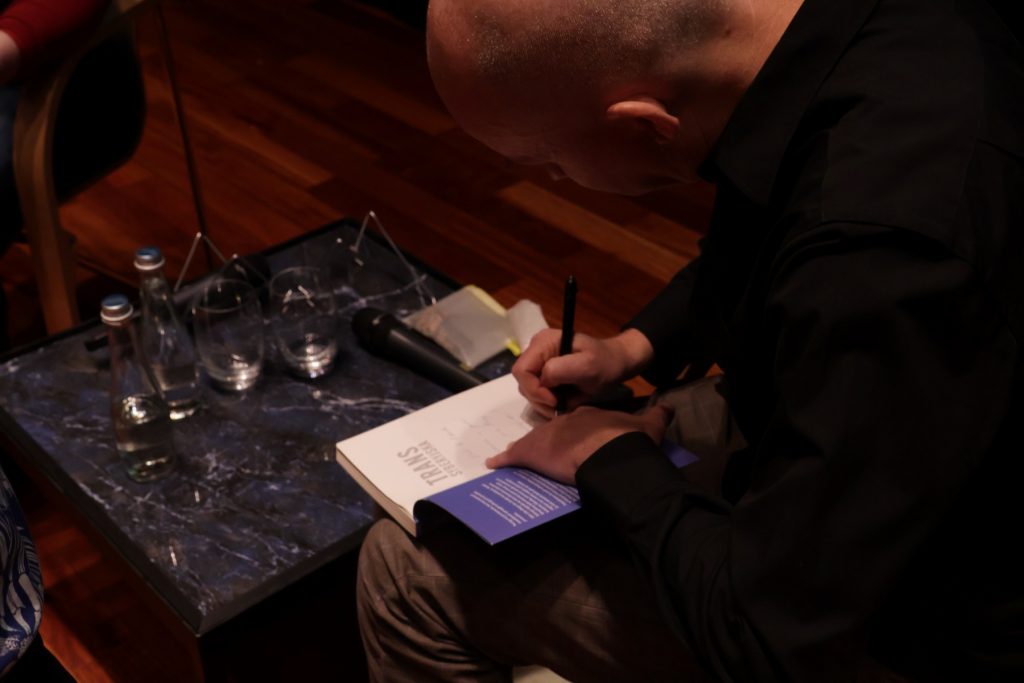 Mężczyzna podpisuje książkę na kolanie, przy stoliku z wodą