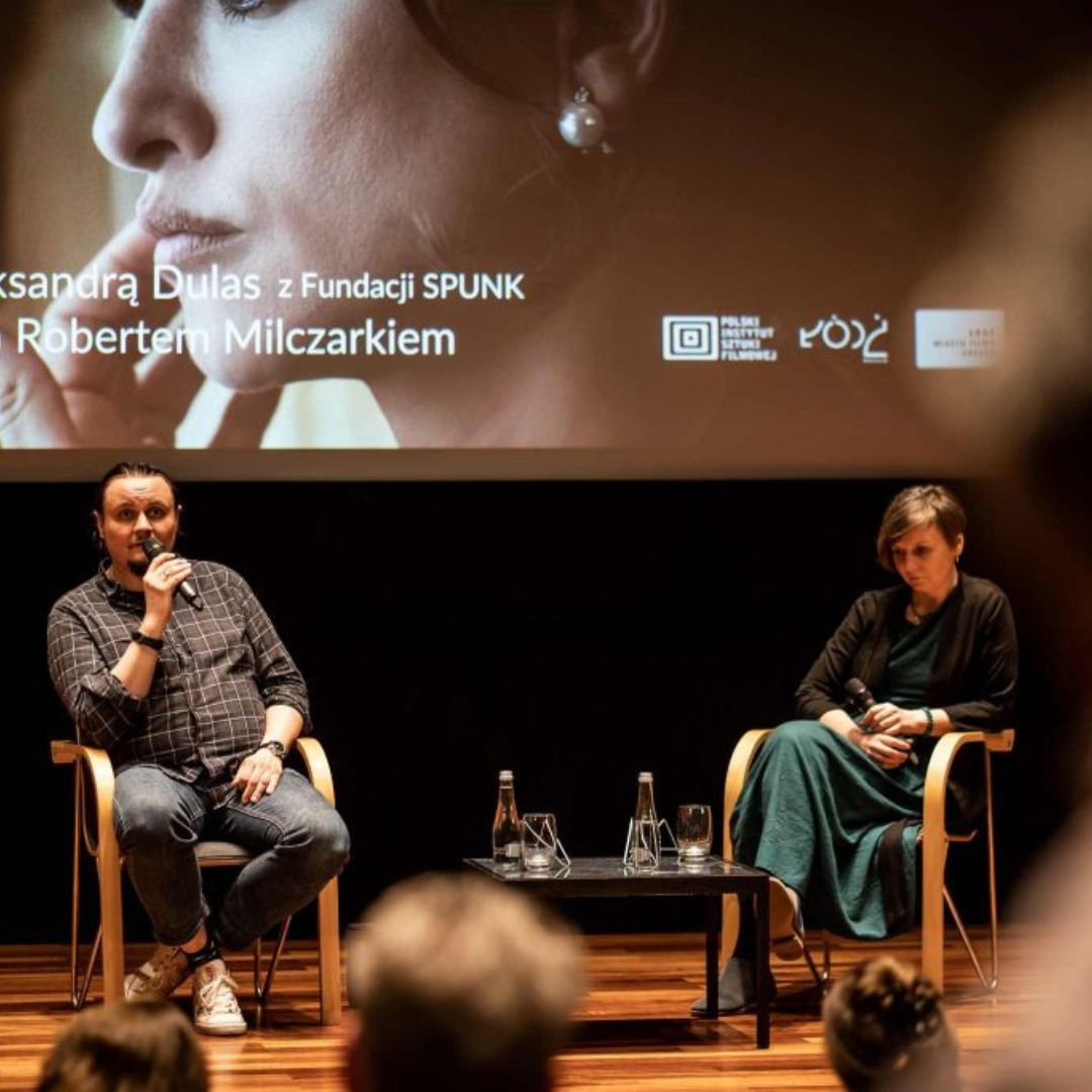 Scena. Od lewej mężczyzna z mikrofonem, obok zamyślona kobieta. Między nimi stolik z wodą