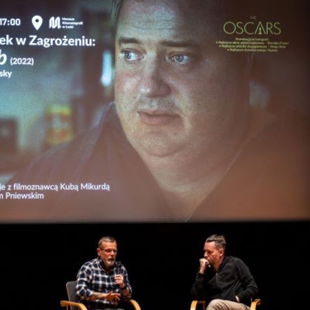 Dwóch mężczyzn siedzi na krzesłach i rozmawiają ze sobą za pomocą mikrofonów. W tle duży, kinowy ekran ze zdjeciem otyłego mężczyzny