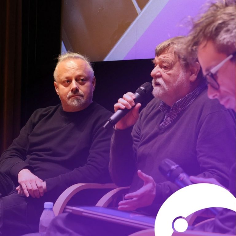 Trzech mężczyzn siedzi na krzesłach i rozmawiają ze sobą. Mężczyzna po środku mówi przez mikrofon, reszta słucha. Zdjęcie pokryte fioletowym tłem. W prawym dolnym rogu logo festiwalu.